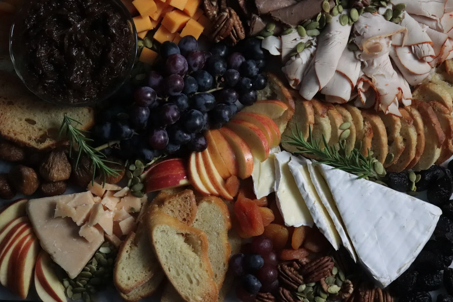 An abundant charcuterie board with cheeses, sliced meats, grapes, apples, toasted bread, nuts and fresh rosemary.