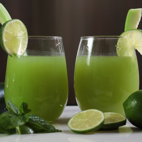 Two glasses filled with cucumber lime agua fresca on a table with limes and cucumber with mint leaves.