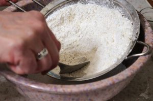 A mixing bowl with powdered sugar and almond flour being combined and pressed through a fine mesh sieve.