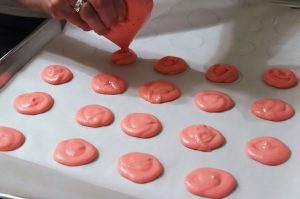 A cooking baking sheet topped with parchment paper with small circular meringue cookies are being piped ready to bake.