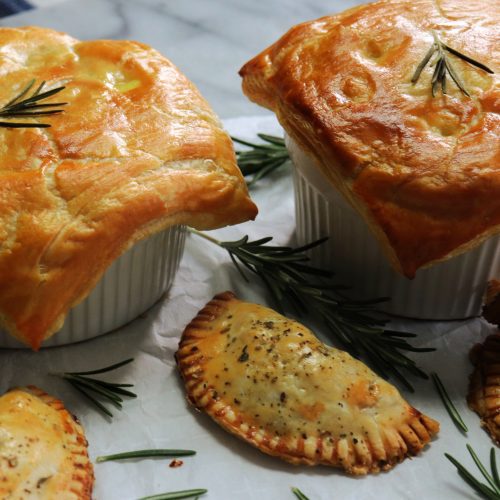 Two kinds of pies made with Thanksgiving leftovers. One is a personal pot pie in a small 8 ounce ramekin. The others are cute little hand pies. They are on a marble board with sprigs of rosemary on parchment paper.
