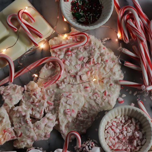 Peppermint candy canes broken into bits and mixed with white chocolate make peppermint bark, in this photo displayed on a marble slab with candy, sprinkles, white chocolate and tiny little Christmas lights.