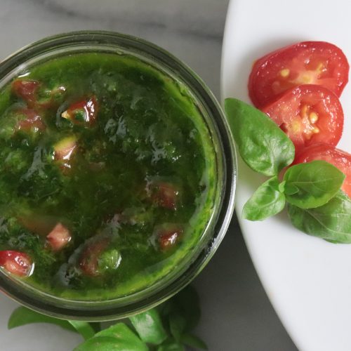 Small bowl of bright green pistou made with fresh basil, garlic, tomato, and olive oil, shown with a spoon and scattered fresh herbs nearby.