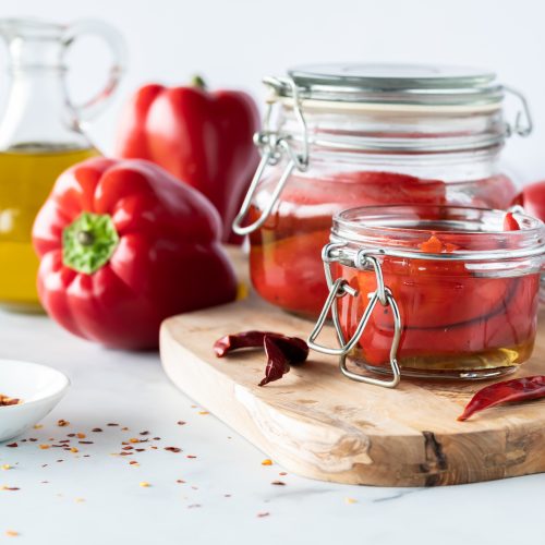 A working area with some fresh red bell peppers on a cutting board and a cruet of olive oil nearby. there are canning jars with roast red peppers inside looking delicious. This post is about how to roast red peppers yourself instead of paying for expensive store bought red peppers.