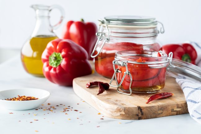 A working area with some fresh red bell peppers on a cutting board and a cruet of olive oil nearby. there are canning jars with roast red peppers inside looking delicious. This post is about how to roast red peppers yourself instead of paying for expensive store bought red peppers.