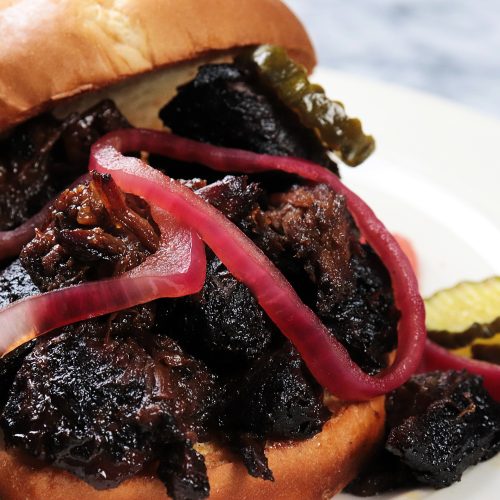 Delicious Poor Man’s Burnt Ends piled high on a warm toasted bun, topped with tangy pickled onions and crisp bread-and-butter pickles, served on a plate for the perfect BBQ bite.