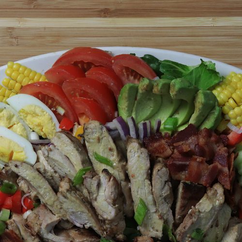 Colorful salad with avocado, tomato, hard boiled eggs, bacon, chopped veggies, grilled corn, and honey mustard chicken, beautifully arranged on a large plate.