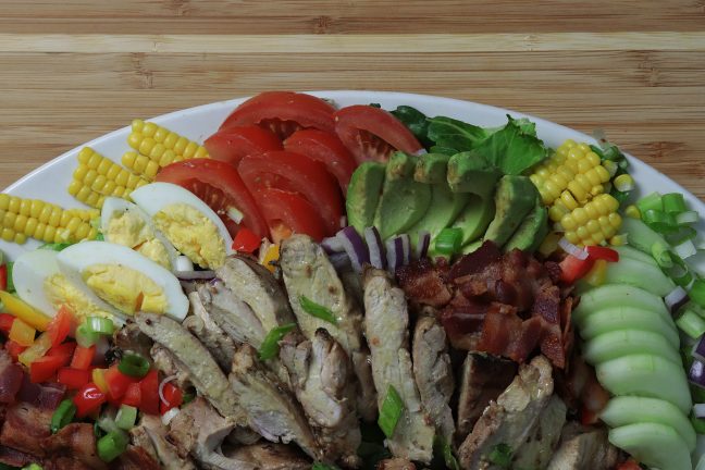 Colorful salad with avocado, tomato, hard boiled eggs, bacon, chopped veggies, grilled corn, and honey mustard chicken, beautifully arranged on a large plate.