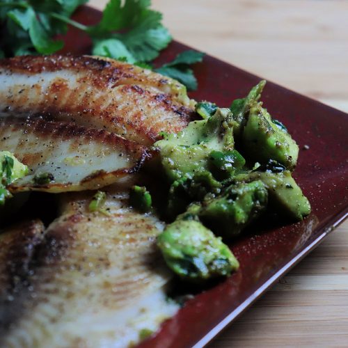 A plate of golden pan-seared tilapia topped with chunky avocado salsa, garnished with cilantro and lime. Served with brown rice and steamed green vegetables.