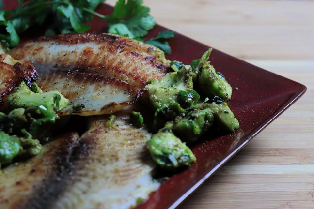 A plate of golden pan-seared tilapia topped with chunky avocado salsa, garnished with cilantro and lime. Served with brown rice and steamed green vegetables.