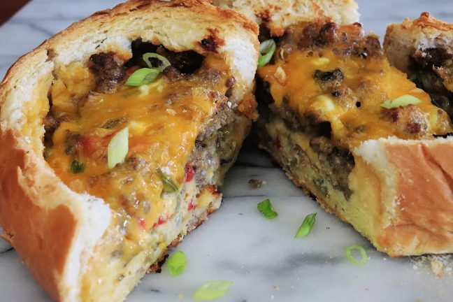 Close-up of a sliced breakfast boat with melty cheese, savory sausage, and sautéed veggies, nestled inside golden, crispy French bread. The warm, gooey filling is perfect for a hearty brunch.