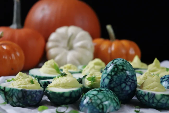 Halloween deviled eggs with deep teal spider-web marbled whites, piped yellow yolk filling, and green onion garnish, arranged on a white platter with orange and white pumpkins behind.
