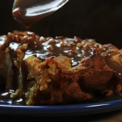 Close-up of golden-brown pumpkin bread pudding with spiced crusty edges on a dark blue plate, pecan praline sauce being drizzled from a spoon.
