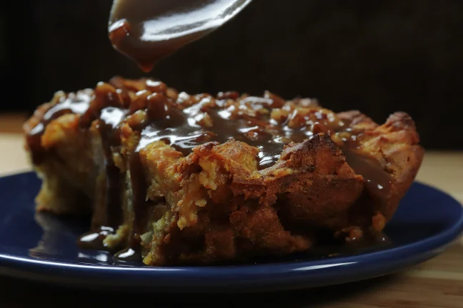 Close-up of golden-brown pumpkin bread pudding with spiced crusty edges on a dark blue plate, pecan praline sauce being drizzled from a spoon.