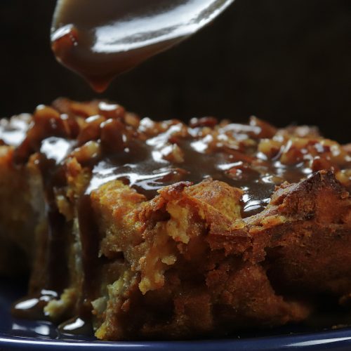 A slice of pumpkin bread pudding on a dark blue plate, glossy pecan praline sauce dripping from a spoon over the top.