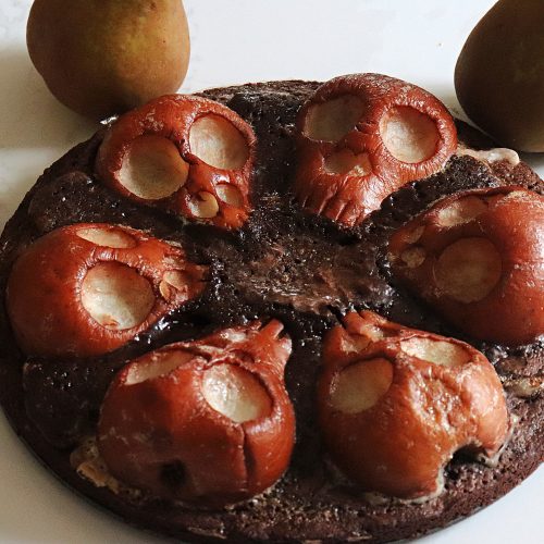 A chocolate brownie cake decorated with pear halves carved into skulls for Halloween, with whole ripe Bosc pears beside it on white marble.