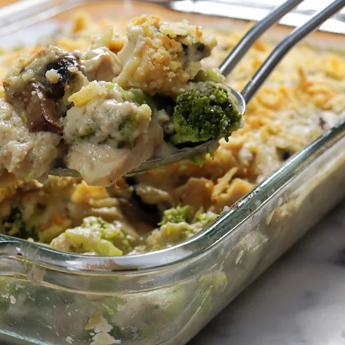 A serving spoon lifting turkey divan casserole with creamy mushroom cheese sauce, broccoli florets, and a golden cheddar topping from a glass baking dish.