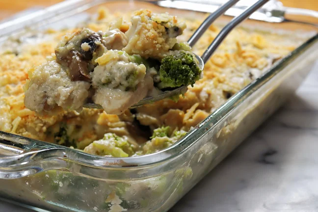 A serving spoon lifting turkey divan casserole with creamy mushroom cheese sauce, broccoli florets, and a golden cheddar topping from a glass baking dish.